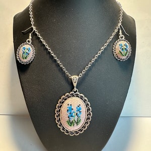 May include: A silver chain necklace with a pendant featuring a blue flower embroidered on a pink background. The pendant is surrounded by a silver frame with a black border. The necklace comes with matching earrings.