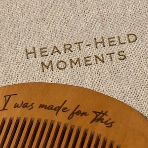 May include: A wooden comb with fine teeth, engraved with the phrase "I was made for this." The comb is set against a beige linen background with the words "HEART-HELD MOMENTS" printed on it. The comb is a light brown color.