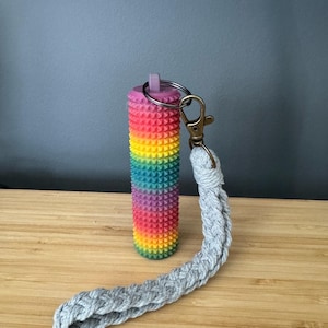 May include: A cylindrical sensory tool with a textured, spiky surface featuring a vibrant rainbow gradient of colors. It is attached to a silver-toned metal split ring and a bronze-colored lobster clasp, which connects to a braided light gray cord.