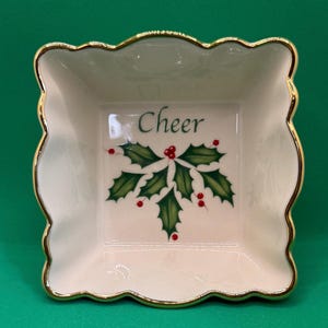 Puede incluir: Un plato cuadrado de cerámica blanquecina con borde festoneado y ribete dorado. El centro presenta la palabra "Cheer" sobre un diseño de acebo y bayas. El plato es probablemente para fines decorativos o de servicio.