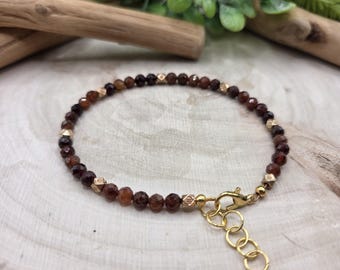 May include: A delicate bracelet featuring small, round, dark red beads interspersed with gold-colored accents. The bracelet has a gold-colored clasp and adjustable chain, and is displayed on a light-colored wooden surface.