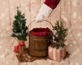 May include: A wooden bucket with a red fabric lining, surrounded by Christmas decorations, including a small Christmas tree, a gift box with red and white stripes, and a small green tree with red berries. The bucket is being held by a hand wearing a white glove.