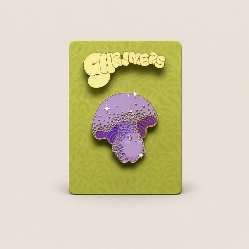 May include: A purple mushroom enamel pin with a smiling face and sparkling details. The pin is on a green card with the word "SHRIMERS" in gold lettering. The pin is approximately 1.5 inches tall.