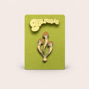 May include: A pin featuring three stylized mushrooms with tan caps and stems, set against a green background with the word "SHRIMERS" in a yellow, whimsical font. The pin is likely a decorative accessory.