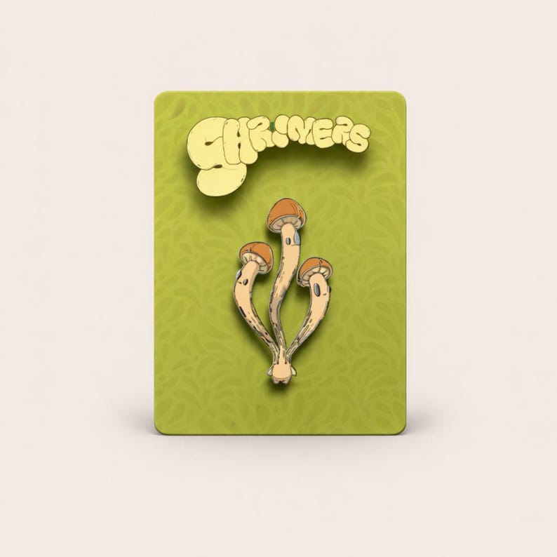 May include: A pin featuring three stylized mushrooms with tan caps and stems, set against a green background with the word "SHRIMERS" in a yellow, whimsical font. The pin is likely a decorative accessory.
