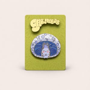 May include: A whimsical enamel pin featuring a cartoon character with blue hair and a smiling face, set against a light blue and white background. The pin is on a green card with the word "SHRIMERS" in gold lettering.