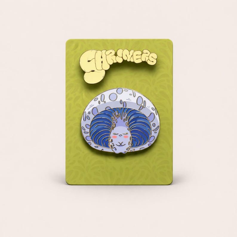 May include: A whimsical enamel pin featuring a cartoon character with blue hair and a smiling face, set against a light blue and white background. The pin is on a green card with the word "SHRIMERS" in gold lettering.