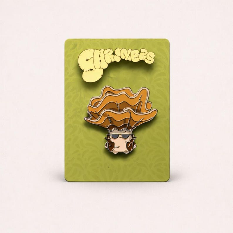 May include: A gold-colored enamel pin featuring a cartoon character with layered orange hair and sunglasses. The pin is on a green card with the word "Charmers" in gold lettering. The pin is a fun, eye-catching accessory.