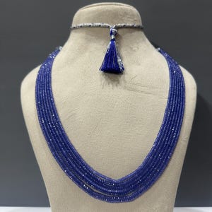 Könnte beinhalten: Eine mehrsträngige Halskette mit mehreren Reihen kleiner, facettierter blauer Perlen. Die Kette verfügt über ein silberfarbenes Choker-Band am oberen Teil, das mit einer kleinen, dunkelblauen Stoffquaste in der Mitte verziert ist.