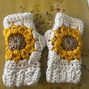 May include: A pair of handmade crochet fingerless gloves. Each glove features a yellow and brown sunflower design on the back of the hand, with a cream-colored cuff and palm. The gloves are made with a textured stitch.
