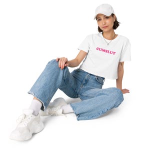 May include: White crop top with the word "CUMSLUT" in pink lettering. The person is wearing light blue jeans, white trainers, and a white baseball cap. The person is sitting on a white surface.