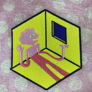 May include: An embroidered patch of a pink figure with blue eyes standing in a yellow room with a blue window. The figure has long arms and legs that extend outside the room. The room is a cube with black outlines.