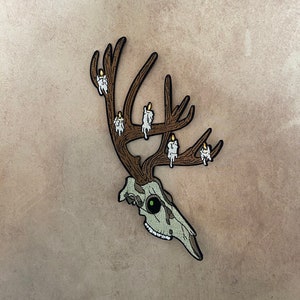 May include: A white deer skull with antlers and lit candles on the antlers. The skull has a green eye and is on a brown background.