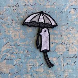 May include: A white and black embroidered patch of a small, cartoon-like figure holding an umbrella. The figure is white with black outlines and the umbrella is black with white stripes.