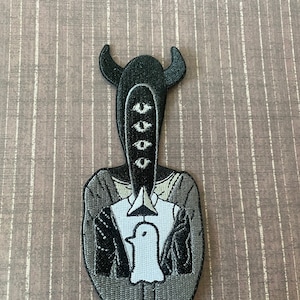 May include: Black and white embroidered patch of a figure with horns, three eyes, and a white bird in its hands.