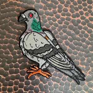 May include: Embroidered iron-on patch of a gray pigeon with red eyes, green feathers, and orange feet. The pigeon is holding a cigarette in its beak.
