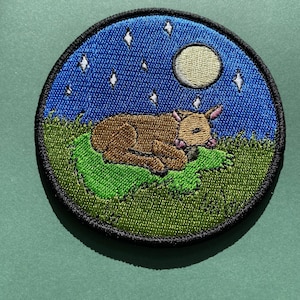 May include: Embroidered patch featuring a brown cow sleeping on a green patch of grass under a blue night sky with a white moon and white stars.