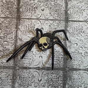 May include: An embroidered patch featuring a black spider with eight long legs and a central body shaped like a human skull. The skull is rendered in shades of gold and black, while the spider's legs and abdomen are solid black thread.