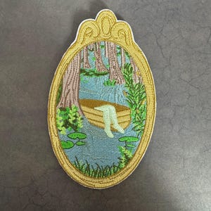 May include: An embroidered patch depicting a person in a boat on a lake, surrounded by trees and lily pads. The patch is framed in a gold oval with a decorative top.