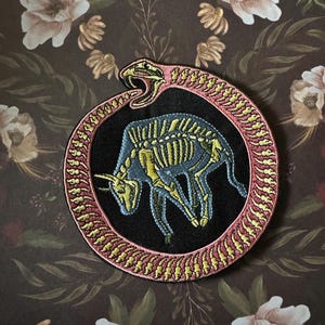 May include: Embroidered patch featuring a blue bull skeleton within a snake biting its tail. The snake is pink and yellow, encircling the bull on a black background. The patch is round and has a decorative border.