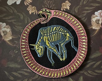 May include: Embroidered patch featuring a blue bull skeleton within a snake biting its tail. The snake is pink and yellow, encircling the bull on a black background. The patch is round and has a decorative border.