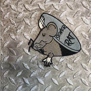May include: A gray and brown embroidered patch of a rat holding a sewer cover with the text "Sewer Rat" on it.