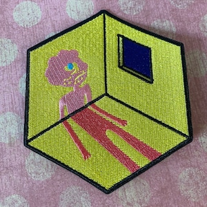 May include: An embroidered patch featuring a pink humanoid figure with a blue eye standing in a yellow room with a blue window. The figure is outlined in red and the room is outlined in black.