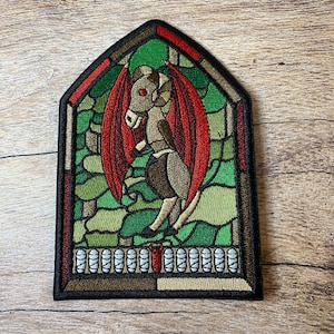May include: Embroidered patch featuring a stained glass window design with a red winged creature inside. The creature has a brown body, red wings, and a red eye. The window is framed in brown and black.