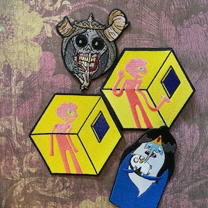May include: Four embroidered patches featuring characters from the animated television series Adventure Time. The patches are yellow, pink, blue, and gray. The characters include a pink humanoid figure in a yellow cube, a blue figure with a crown and a white bird, a pink humanoid figure in a yellow cube, and a gray figure with horns and a crown.