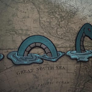 May include: Embroidered patches featuring the Loch Ness Monster in teal, gray, and blue. The patches depict the monster's head and arched back emerging from the water, with a fin visible. Set against a vintage world map background.