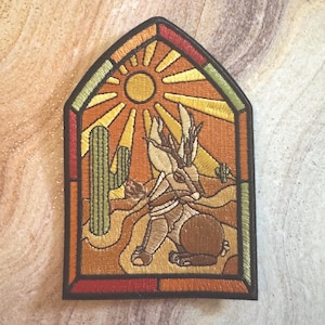 May include: An embroidered patch in the shape of a pointed arch window featuring a jackalope in a desert landscape. The design includes a bright yellow sun, a green cactus, and a brown jackalope with antlers, all outlined in black thread.
