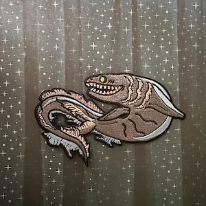 May include: A brown embroidered patch featuring a stylized sea serpent with a toothy grin and green eyes. The patch has a white border and detailed stitching, perfect for adding to clothing or accessories.