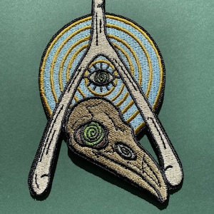 May include: An embroidered patch with a white bone-like structure forming a triangle shape. Inside the triangle is a brown and green skull with a green eye. The triangle is surrounded by a blue and gold circle with concentric rings.