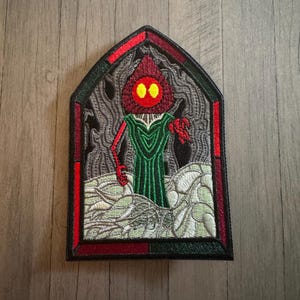 May include: An embroidered patch shaped like a pointed arch window featuring a figure with a red hood and yellow eyes wearing a green robe. The scene includes a dark gray forest background and light green foreground elements, all framed by a black border.