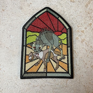 May include: An embroidered patch depicting a stained glass window with a sunset scene. A gray wolf-like creature with red eyes is attacking a brown and white deer. The scene is set against a yellow and orange background.