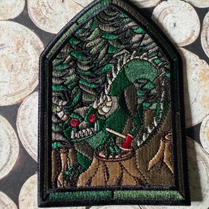 May include: Embroidered patch featuring a green dragon with red eyes, holding a small red axe. The dragon is set against a forest scene with a brown tree stump. The patch has a black border and a gothic arch shape.