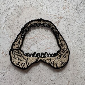 May include: A beige embroidered patch of a shark's jaw with black stitching. The patch is shaped like an open mouth with sharp teeth.