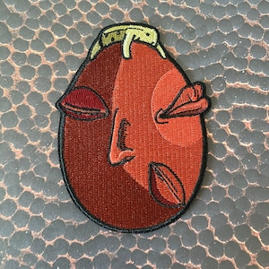 May include: A red embroidered patch with a stylized face with closed eyes and a winking mouth. The face is outlined in black and has a yellow band around the top.