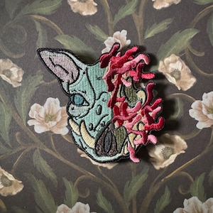 May include: Embroidered patch featuring a cat skull design with a teal and pink color scheme. The patch has a black border and includes red and pink floral elements on the right side. The background is a floral pattern.