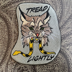 May include: Embroidered patch with a gray background featuring a cartoon cat with green eyes and a yellow and black snake in its mouth. The text "TREAD LIGHTLY" is embroidered below the cat.