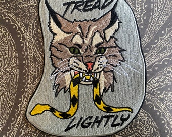May include: Embroidered patch with a gray background featuring a cartoon cat with green eyes and a yellow and black snake in its mouth. The text "TREAD LIGHTLY" is embroidered below the cat.