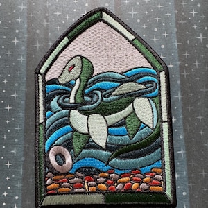 May include: A green and blue embroidered patch depicting a sea serpent with a white body and red eyes, swimming in blue water with white waves. The patch is framed in a green and white border, resembling a stained glass window.
