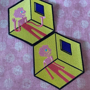 May include: Two embroidered patches featuring a pink figure with a single blue eye inside a yellow cube with a blue window. The figure is in a stretched out pose with long limbs.