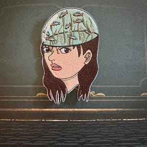 May include: An embroidered patch featuring a person with dark brown hair and a light blue, dome-shaped head. The head is filled with a pattern of small, swimming fish in shades of orange and blue, set against a background of vertical green lines.