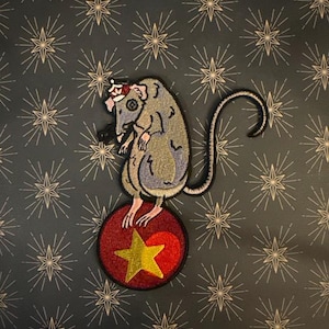 May include: Embroidered patch featuring a rat balancing on a red ball with a gold star. The rat is gray and blue, wearing a crown. The background is dark blue with a starburst pattern.