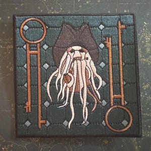 May include: A square embroidered patch featuring a central figure with a tentacled face wearing a dark hat, flanked by two tall, thin copper-colored keys. The background is a dark teal grid pattern with small light blue diamond accents.