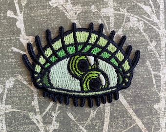 May include: Green embroidered eye patch with black stitching and a white iris. The eye has two pupils.
