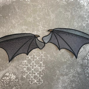 May include: Two black felt bat wings with a gray embroidered design. The wings are on a gray textured background.