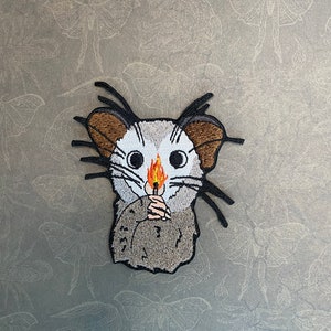 May include: Embroidered patch of a gray possum holding a lit match. The possum has large, white eyes and brown ears. The background is a gray fabric with a pattern of moths.