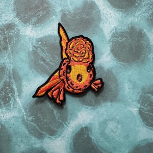 May include: A red and yellow embroidered patch of a goldfish with a rose on its head. The goldfish is swimming to the right.